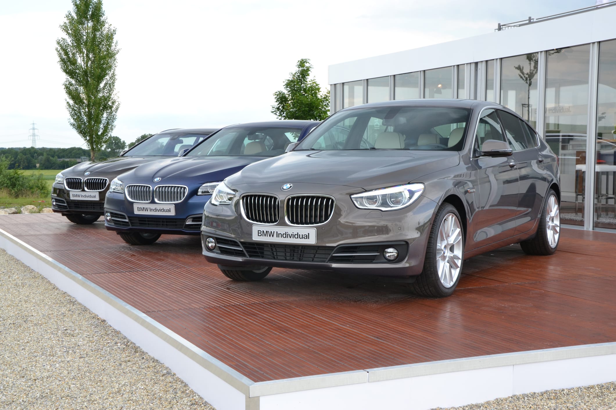 Three sleek, modern BMW cars in different shades of gray are parked on a wooden platform outdoors. The cars exude luxury and sophistication.