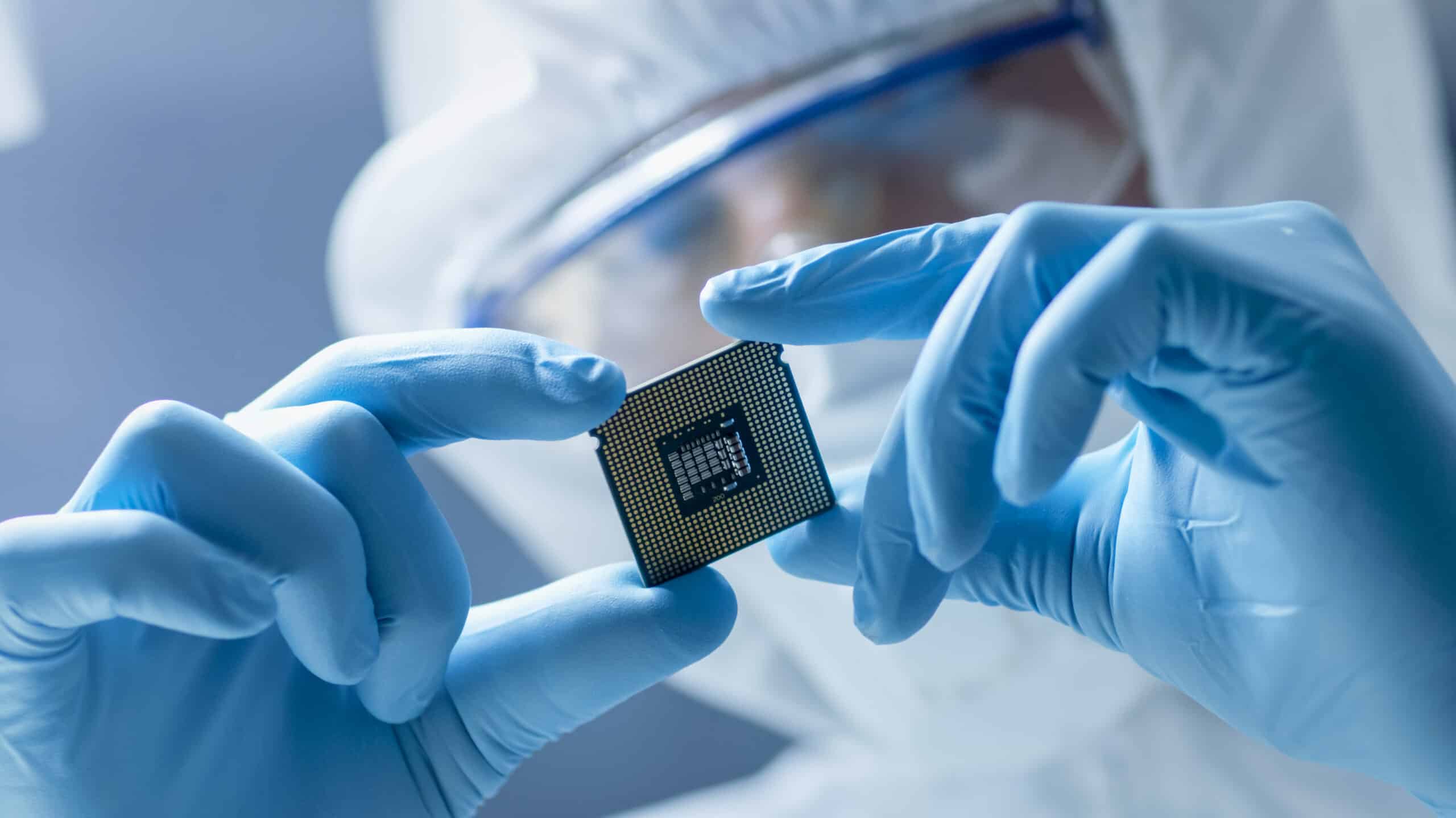 Person in protective gear and gloves holds a microchip delicately, emphasizing precision and technology in a sterile, high-tech environment.
