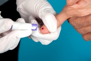 Gloved hands using a lancing device to prick a fingertip with a drop of blood