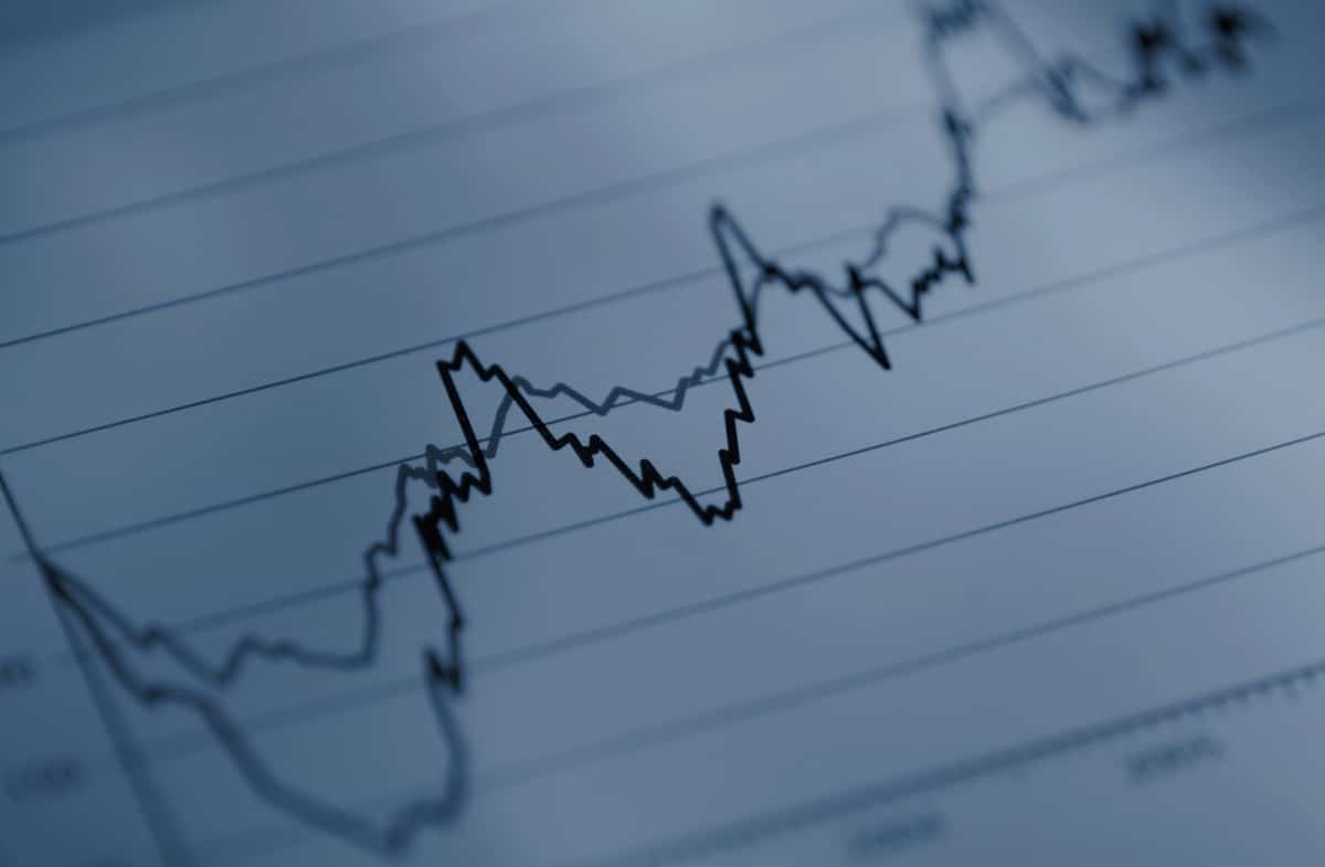 A close-up of a financial line graph with two jagged lines representing data trends moving across a grid, showing rises and falls over time. The image has a blue tone and blurred edges.
