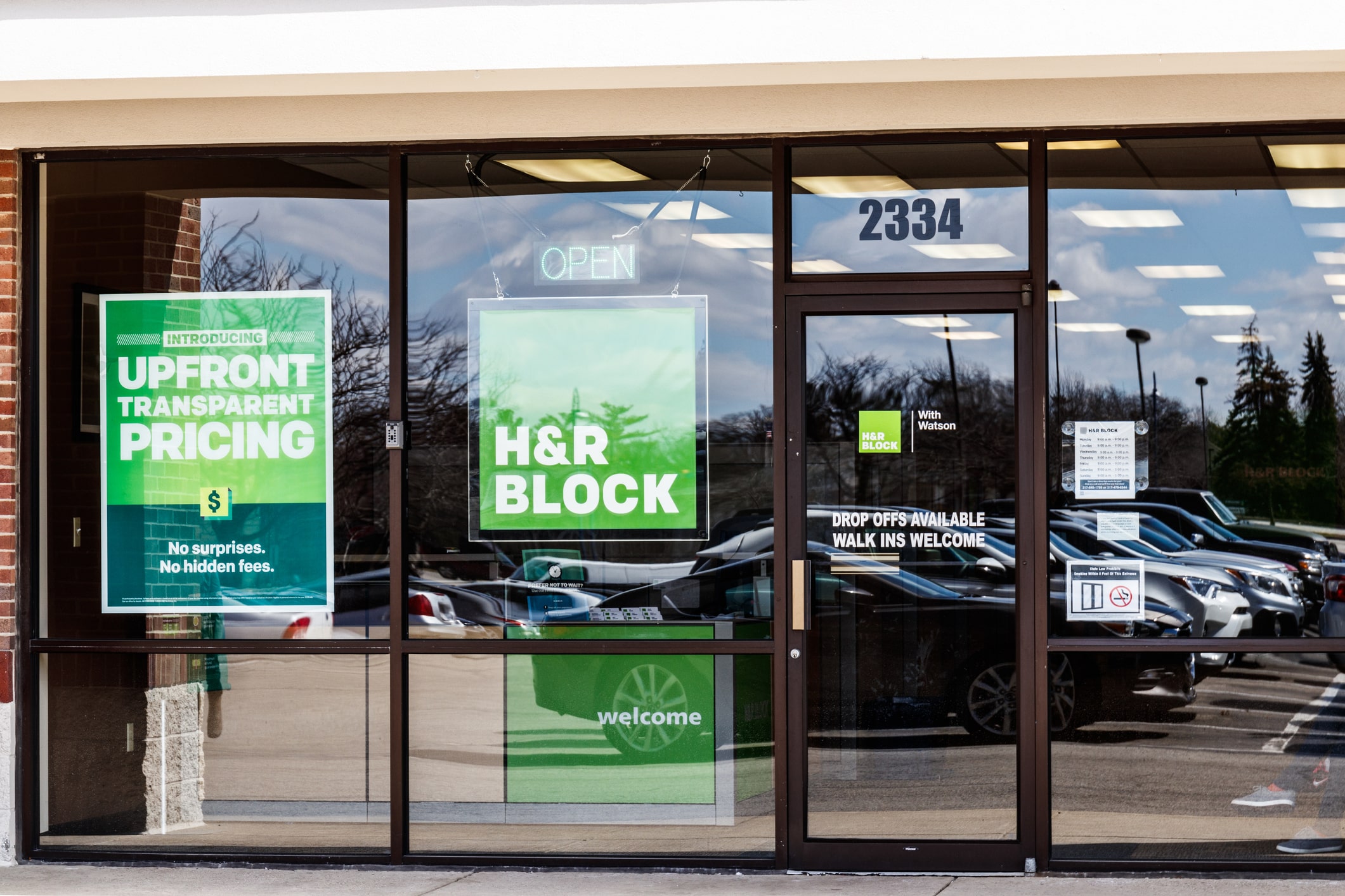 Storefront of an H&R Block office with large green signs advertising upfront transparent pricing. Open sign is lit, and cars reflect in the glass.