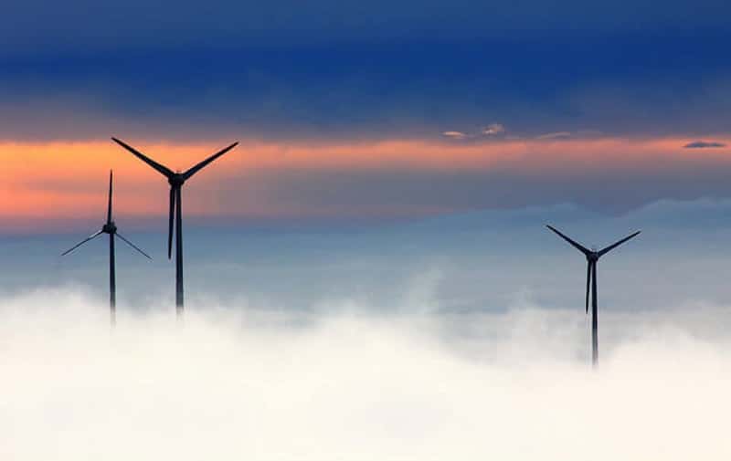 Three wind turbines rise above thick clouds, silhouetted against a vibrant sunset with deep blue and orange hues, conveying a sense of serenity.