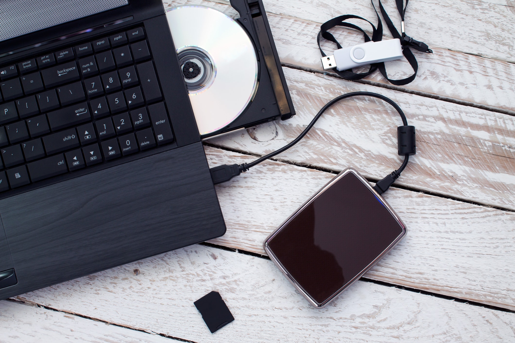 Laptop on a rustic white table with an open CD drive, connected external hard drive, USB flash drive, and SD card. Tech setup conveys connectivity.