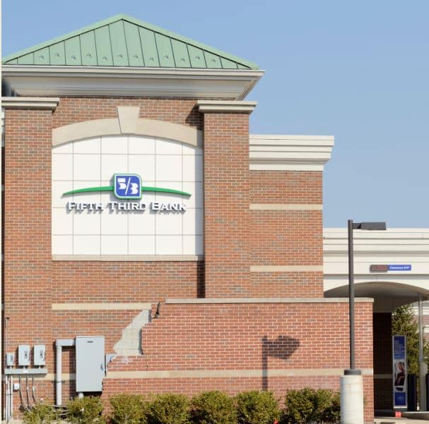 Red brick building with a green roof features a "Fifth Third Bank" logo. Surrounding greenery adds a welcoming touch under a clear blue sky.