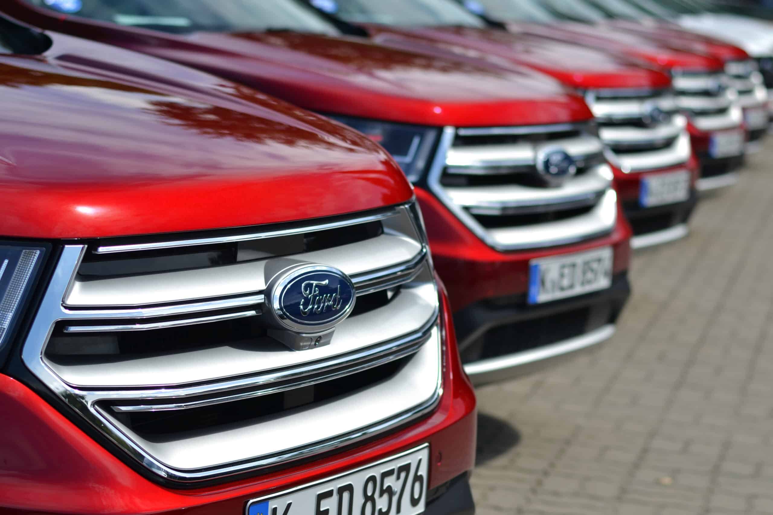 Row of red Ford vehicles parked in a line on a paved surface, focusing on the front grills and license plates.