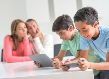 Two boys using a tablet and smartphone with adults blurred in background