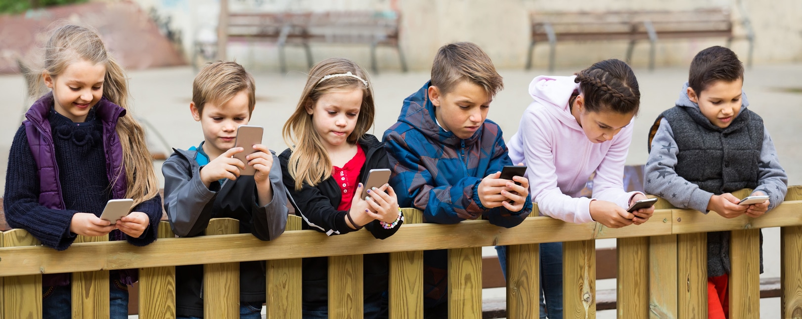 Six children leaning on a wooden fence, all focused on their smartphones, expressing engagement and concentration in an outdoor setting.