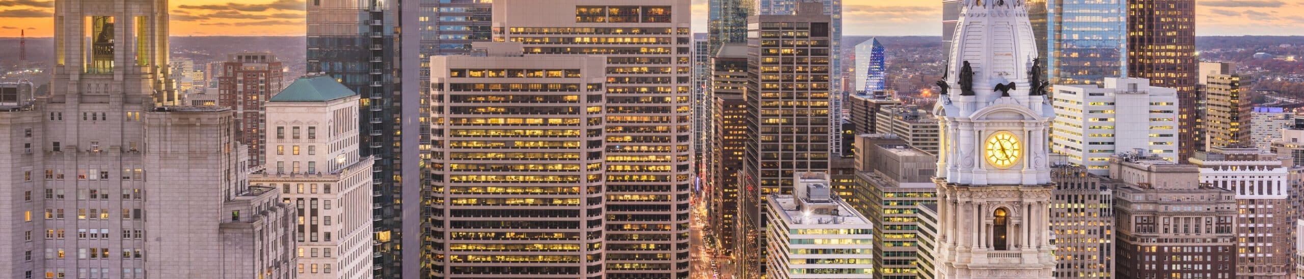 Panoramic view of downtown Philadelphia at dusk with high-rise buildings and the historic City Hall clock tower glowing against the evening sky.