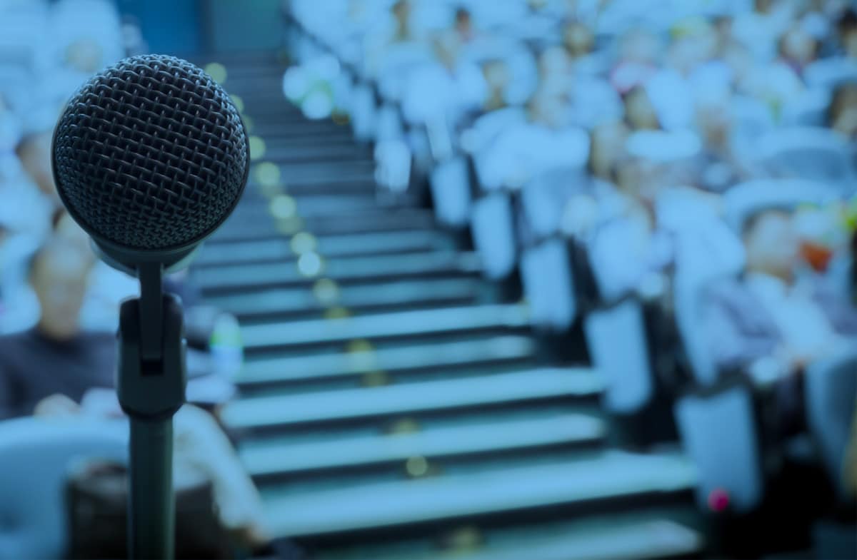 Microphone on a stand in the foreground with a blurred audience seated in an auditorium, suggesting a conference, seminar, or public speaking event.