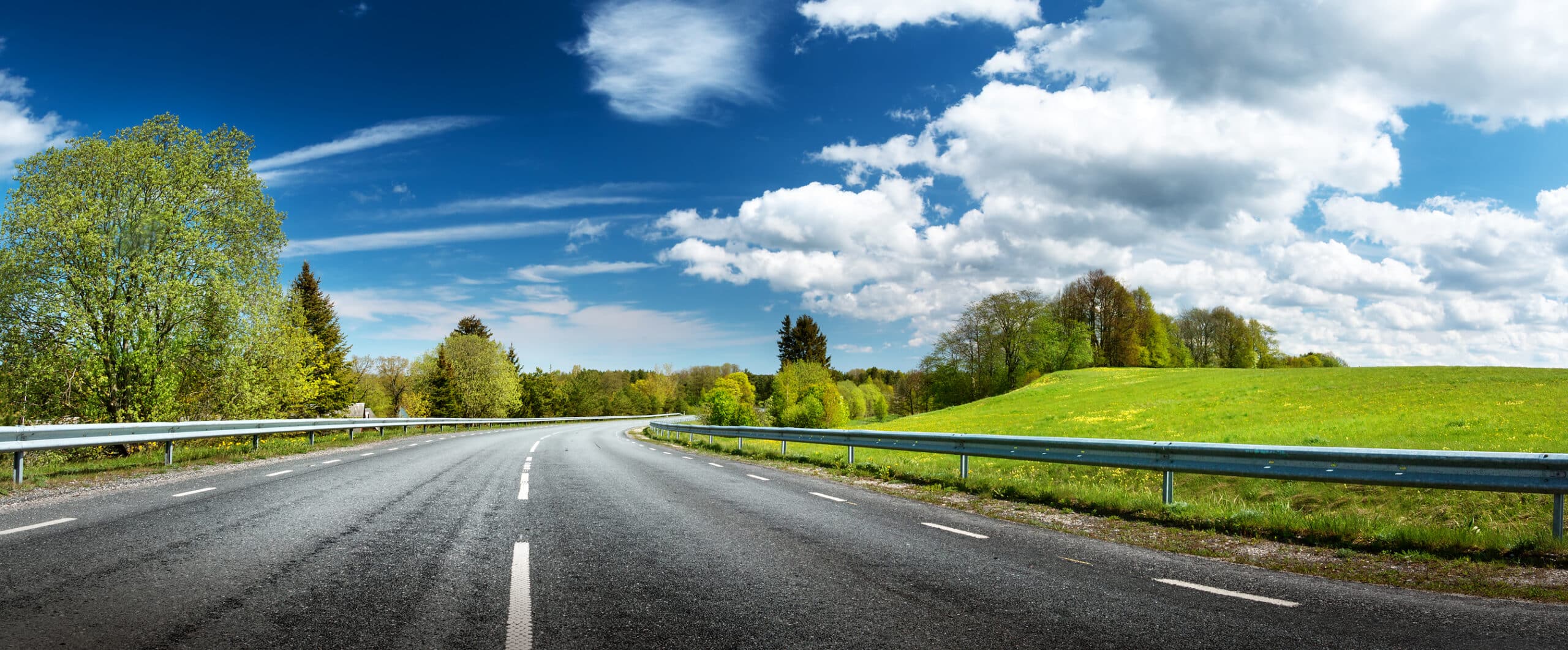 Winding asphalt road with white lines and guardrails, surrounded by green trees and a grassy hill, under a blue sky with white clouds