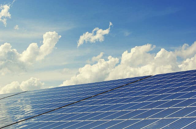 Array of blue solar panels angled upward, filling the bottom half of the image against a cloudy blue sky.